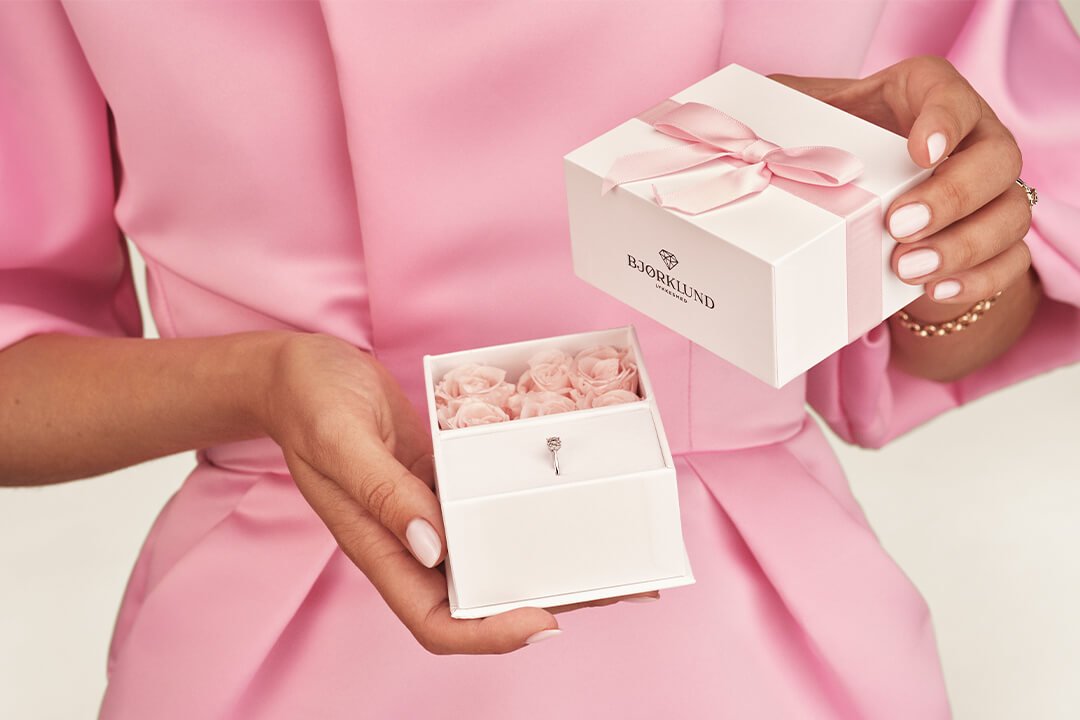 Nærbilde av en gavepakke fra Bjørklund med ring og rosa blomster som holdes av en kvinne i rosa ballkjole.