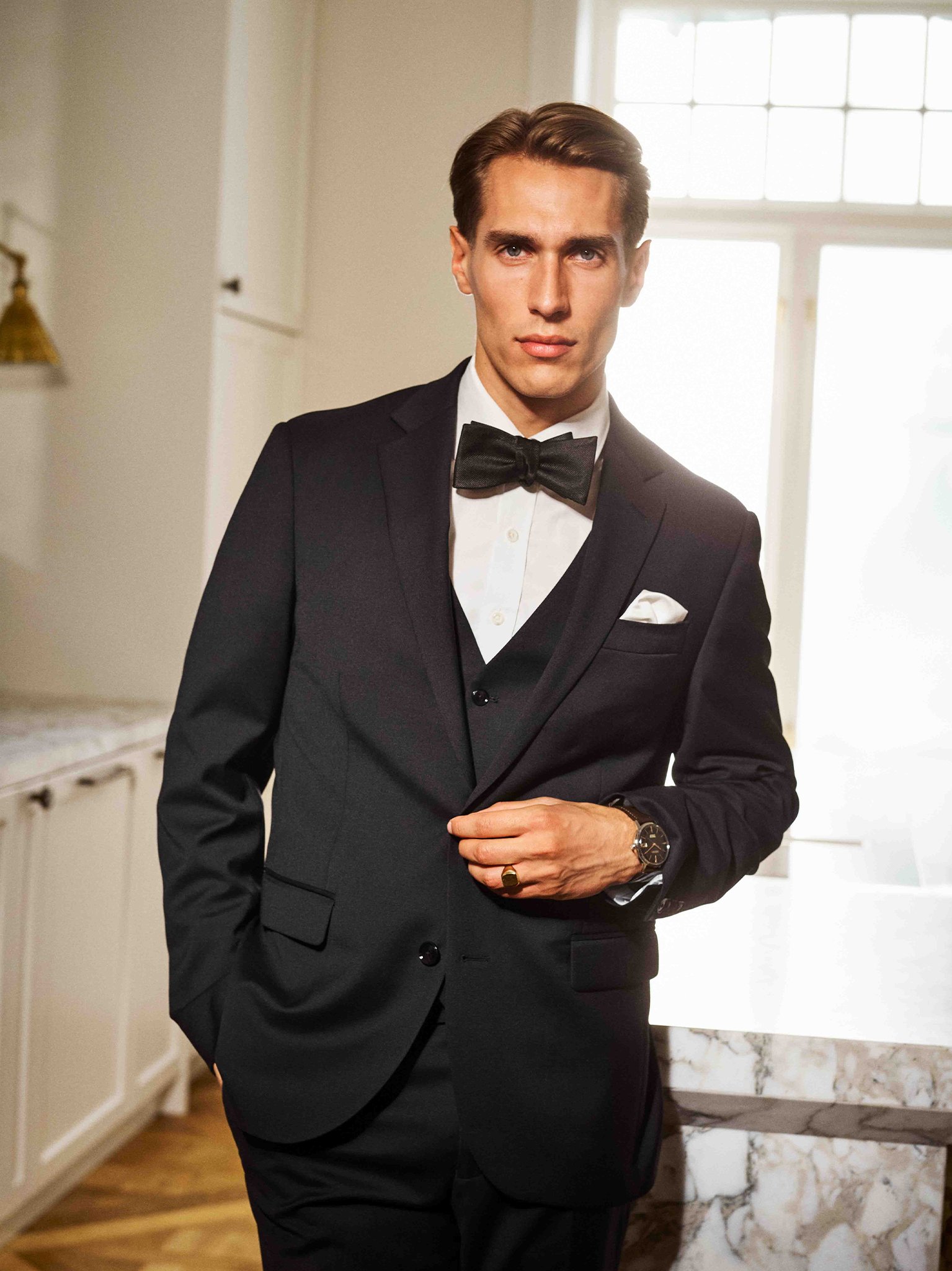 Mann i sort smoking med sløyfe og klokke står ved marmorkamin i lyst rom med store vinduer.