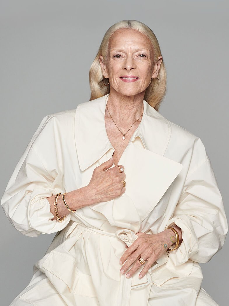 Eldre kvinne i lys kjole står mot grå bakgrunn og bærer flere gullsmykker, inkludert armbånd, ringer og halskjede.