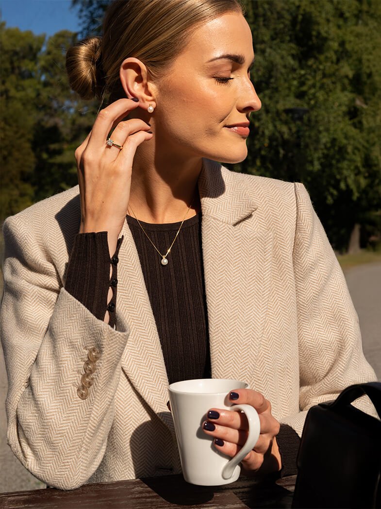 Elegant kvinne med oppsatt hår i beige fishbone-blazer og mørk topp, bærer gullring og øredobber mens hun holder hvit kaffekopp i høstlig sollys