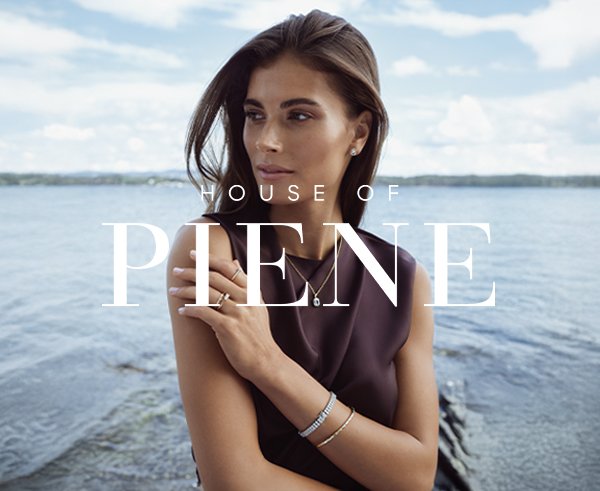 Kvinne står utendørs ved sjøen med smykker på hals, håndledd og fingre, iført mørk topp med vann og himmel i bakgrunnen.