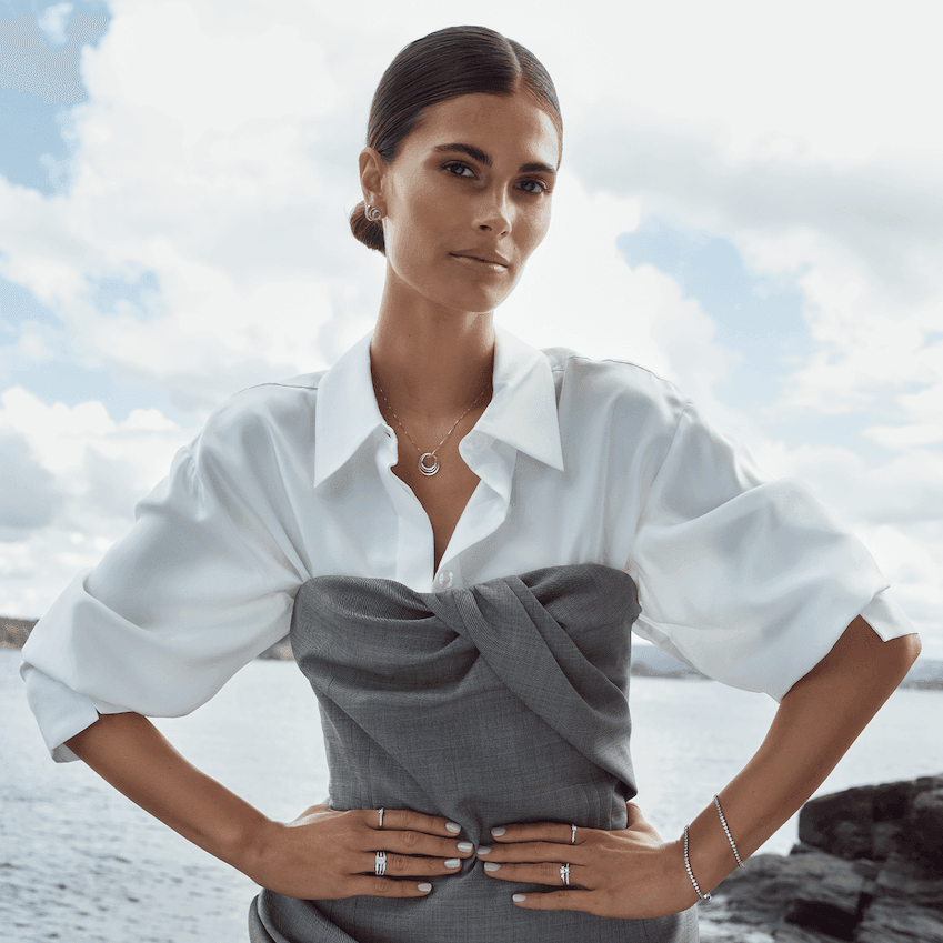 Kvinne med hvit skjorte og grå kjole ved sjøen, iført gullsmykker og diamantringer