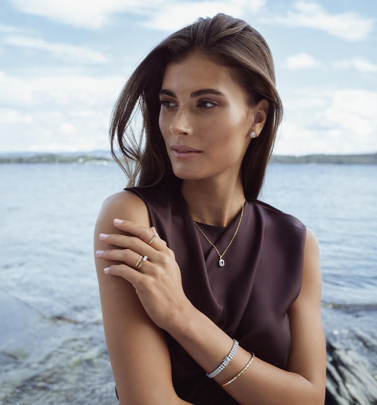 Kvinne med langt hår står ved sjøen iført mørk topp, smykker, ringer, armbånd og øredobber i gull og sølv.