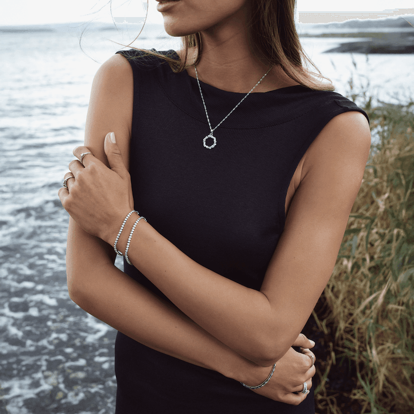 Kvinne ved sjøen kledd i mørk ermeløs topp, med halskjede, armbånd og ringer i sølvfarget metall.