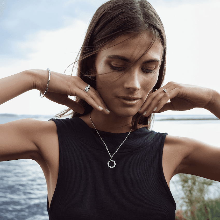Kvinne ved sjøen med mørkt hår, iført mørk ermeløs topp og smykker i sølvfarget metall, inkludert halskjede og ringer.