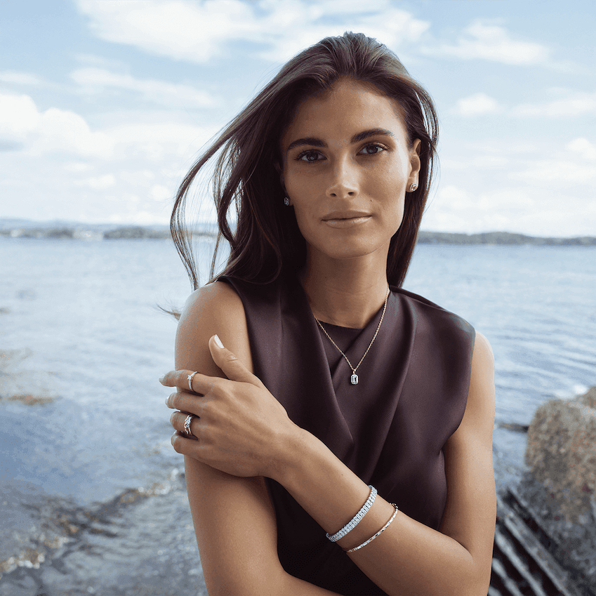 Kvinne med langt hår står ved sjøen og bærer smykker som halskjede, armbånd, ringer og øredobber i sølvfarget metall.