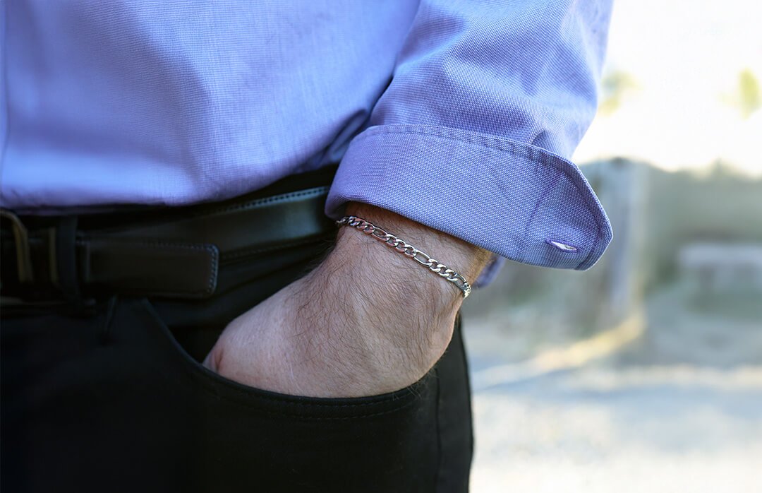 Armbånd til herre En mann med lyseblå skjorte og svart bukse har hånden i lommen og bærer et sølvarmbånd.