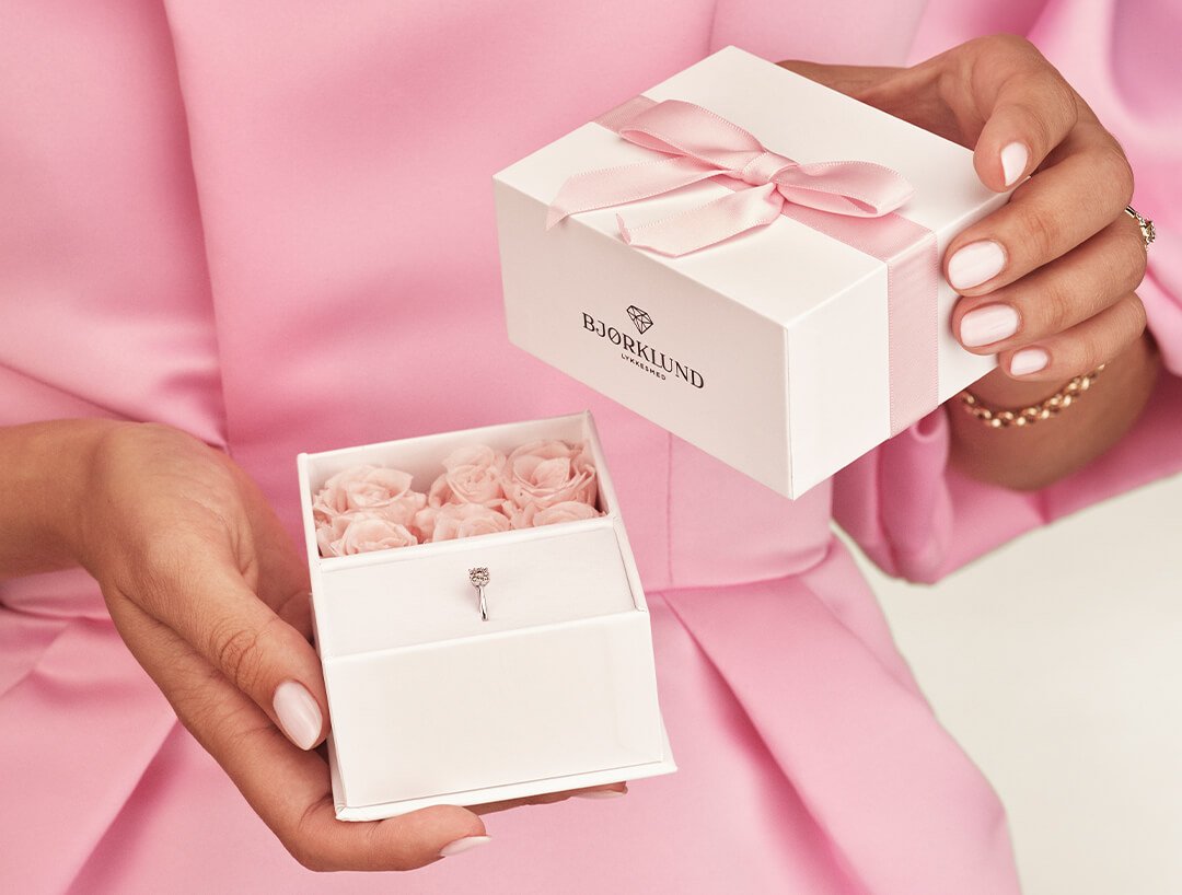 Gaveeske til mamma En kvinne med rosa ballkjole holder en hvit gaveeske med rosa bånd, med rosa blomster og en ring inni.