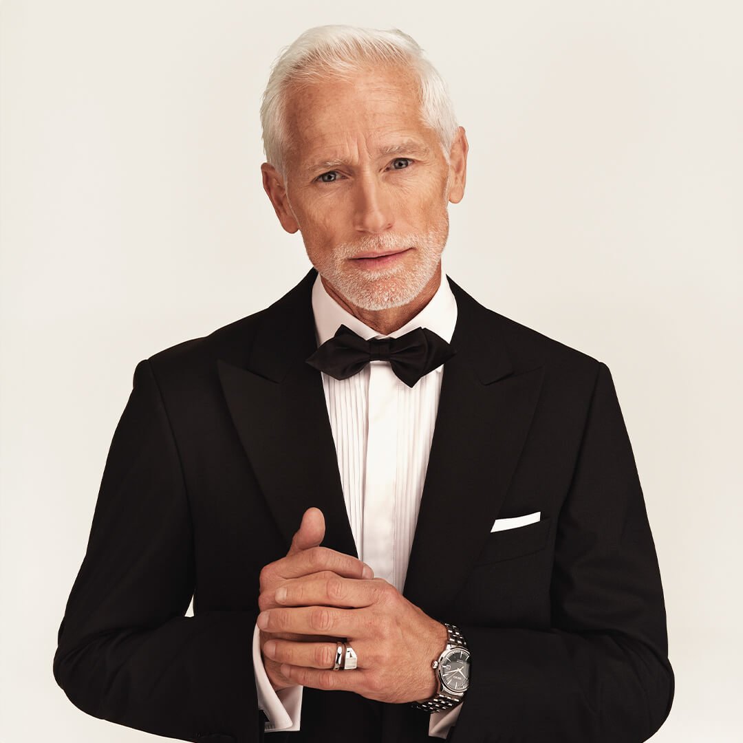 Mann i sort smoking med sløyfe og klokke står ved marmorkamin i lyst rom med store vinduer.