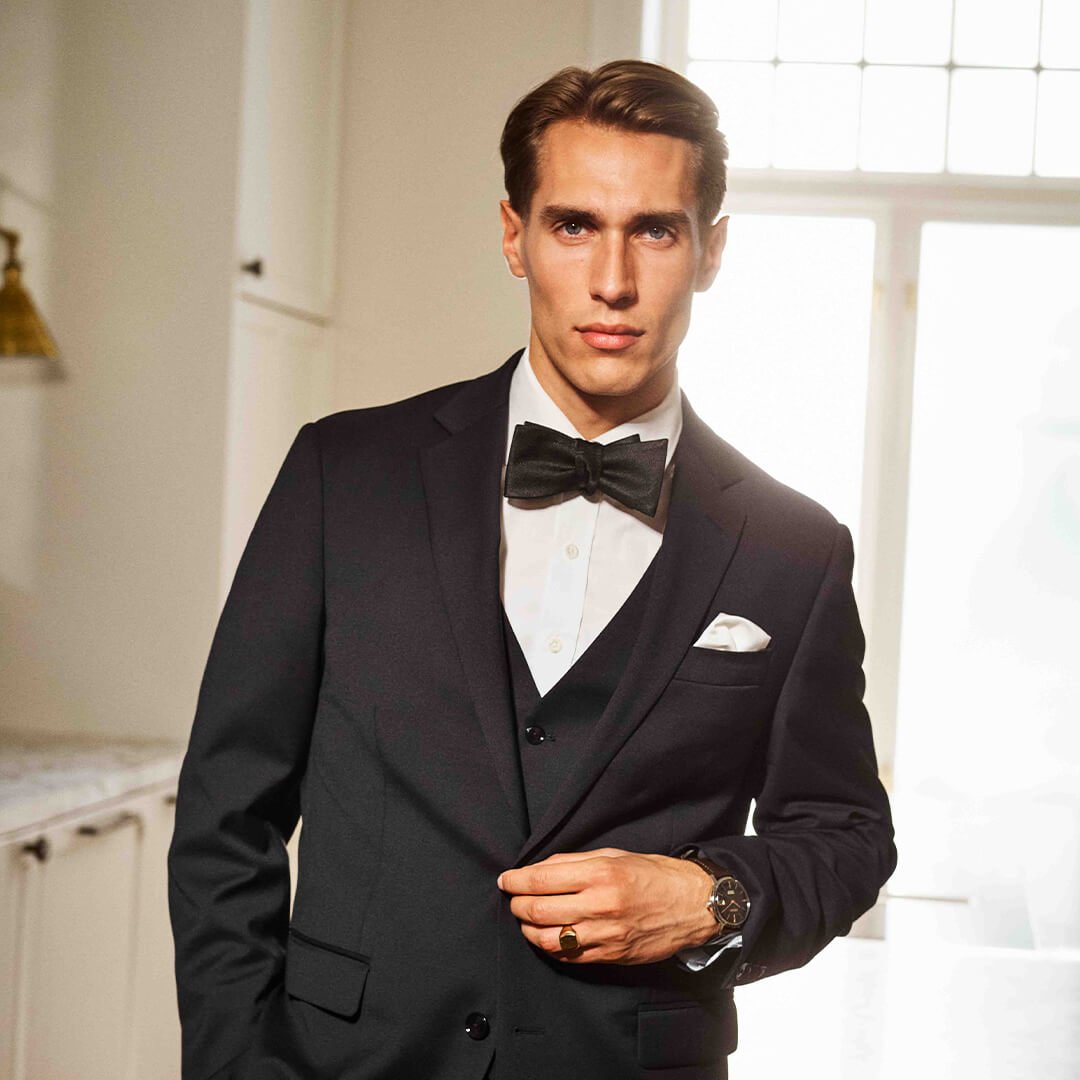 Mann i sort smoking med sløyfe og klokke står ved marmorkamin i lyst rom med store vinduer