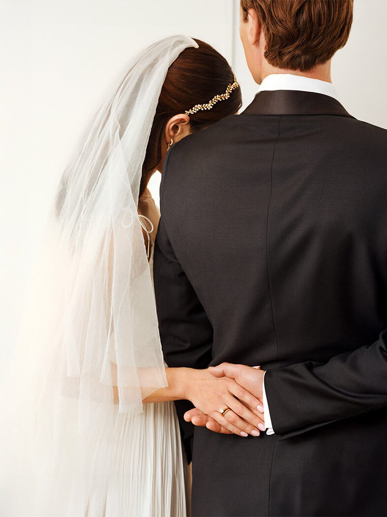 Brudepar sett bakfra, der bruden i slør og kjole holder rundt brudgommens arm, begge bærer gifteringer.