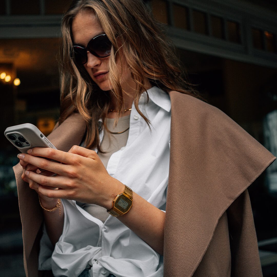 En moteriktig kvinne med langt, bølgete hår står utenfor en kafé og ser på telefonen sin. Hun har på seg en hvit skjorte, en beige kåpe hengende over skuldrene og en gullfarget digital klokke på håndleddet. 