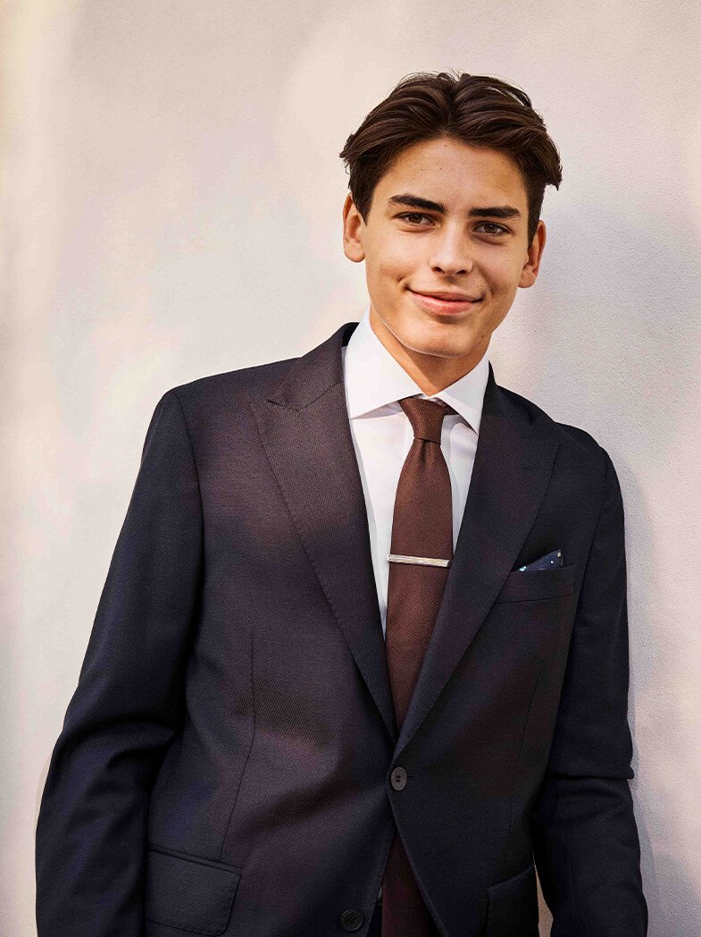 Ung mann med brunt hår i mørk dress, hvit skjorte, brunt slips med slipsnål og mønstret lommetørkle, mot en lys vegg.