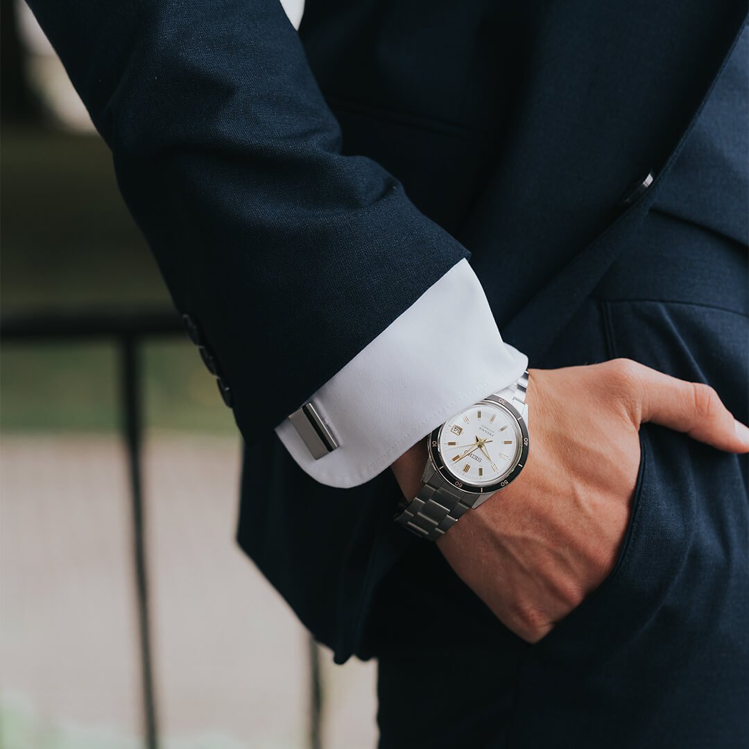 Kjøp klokke til konfirmanten Nært bilde av en ung mann i dress som holder hånden i lommen, med tommelen utenfor. Han har en elegant klokke på håndleddet.