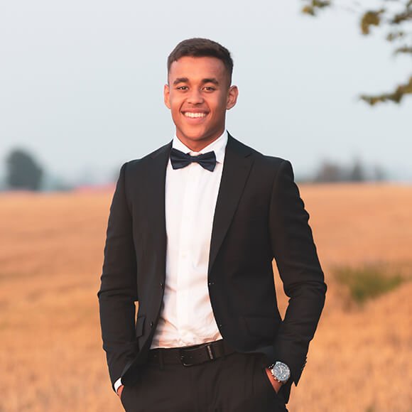 En tenårinings gutt med mørkeblå dress og blå skjorte, som fikser på sitt slips mens han smiler og ser ut til siden.