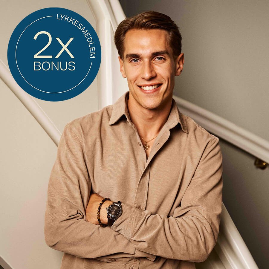 Mann med beige skjorte smiler og har armene i kors, iført armbåndsur, perlearmbånd og kjede med anheng i gull. Sirkelgrafikk med "2x bonus lykkesmedlem"