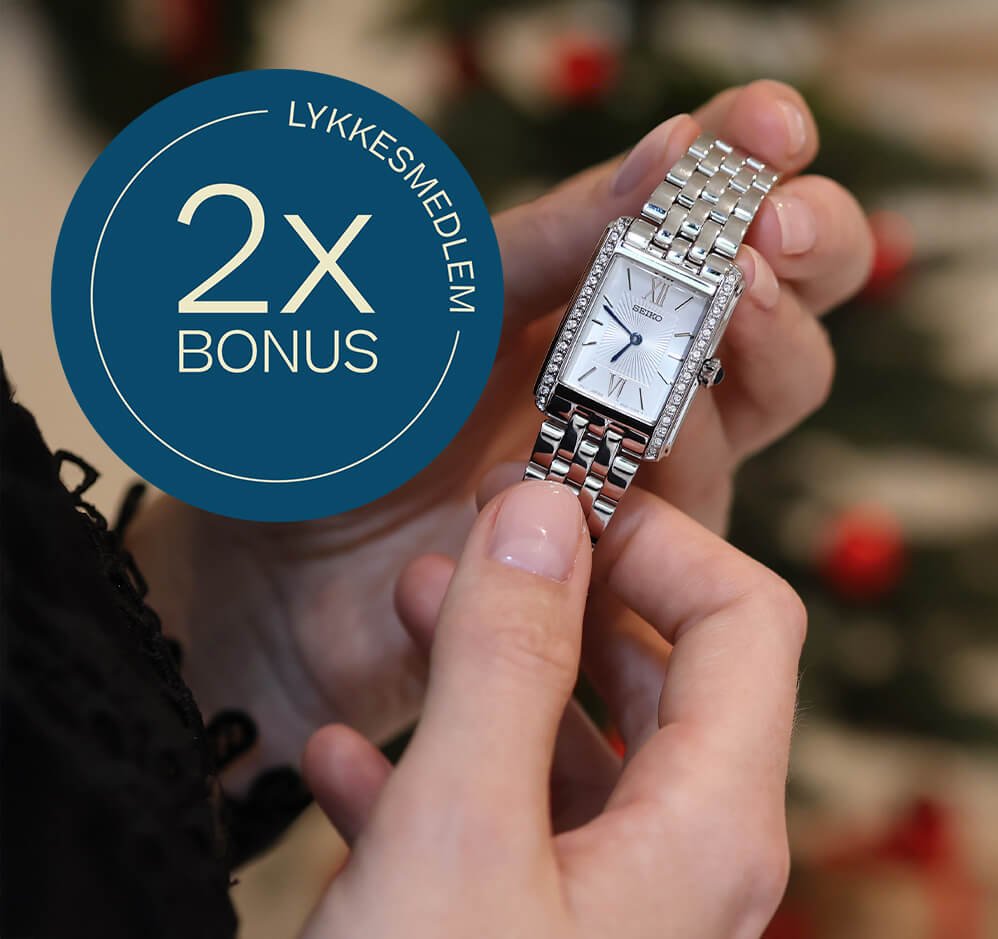 Kvinnehender holder en sølvfarget Seiko-klokke foran juletre, med blå sirkeltekst som viser «Lykkesmedlem 2x bonus».