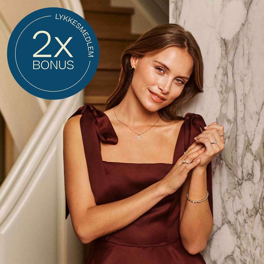Kvinne i vinrød kjole smiler og viser smykker og ringer, med blå sirkeltekst som viser «Lykkesmedlem 2x bonus».