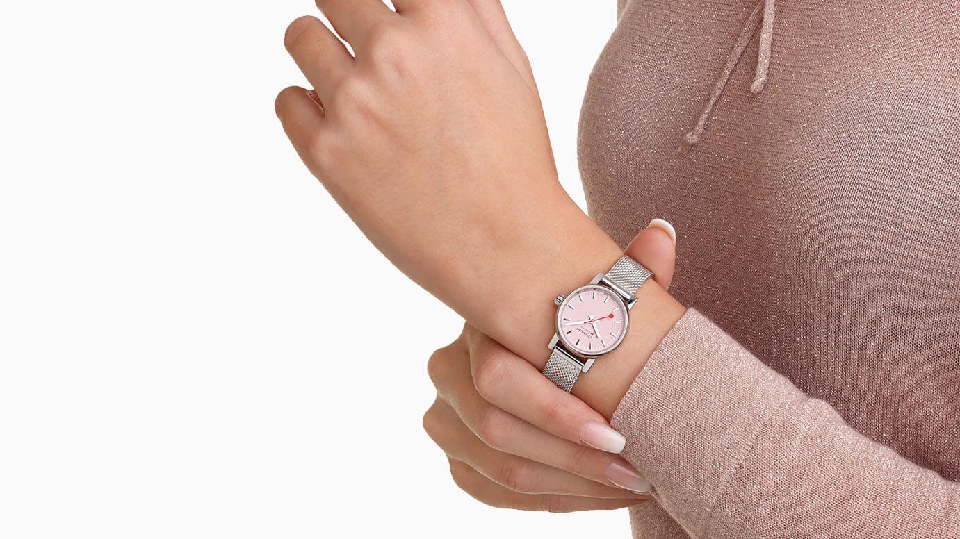 EVO2–kolleksjonen En kvinne i rosa, tynn genser med knyting i halsen, justerer Mondaine-klokken sin i sølv og rosa, som hun har på armen litt før håndleddet.
