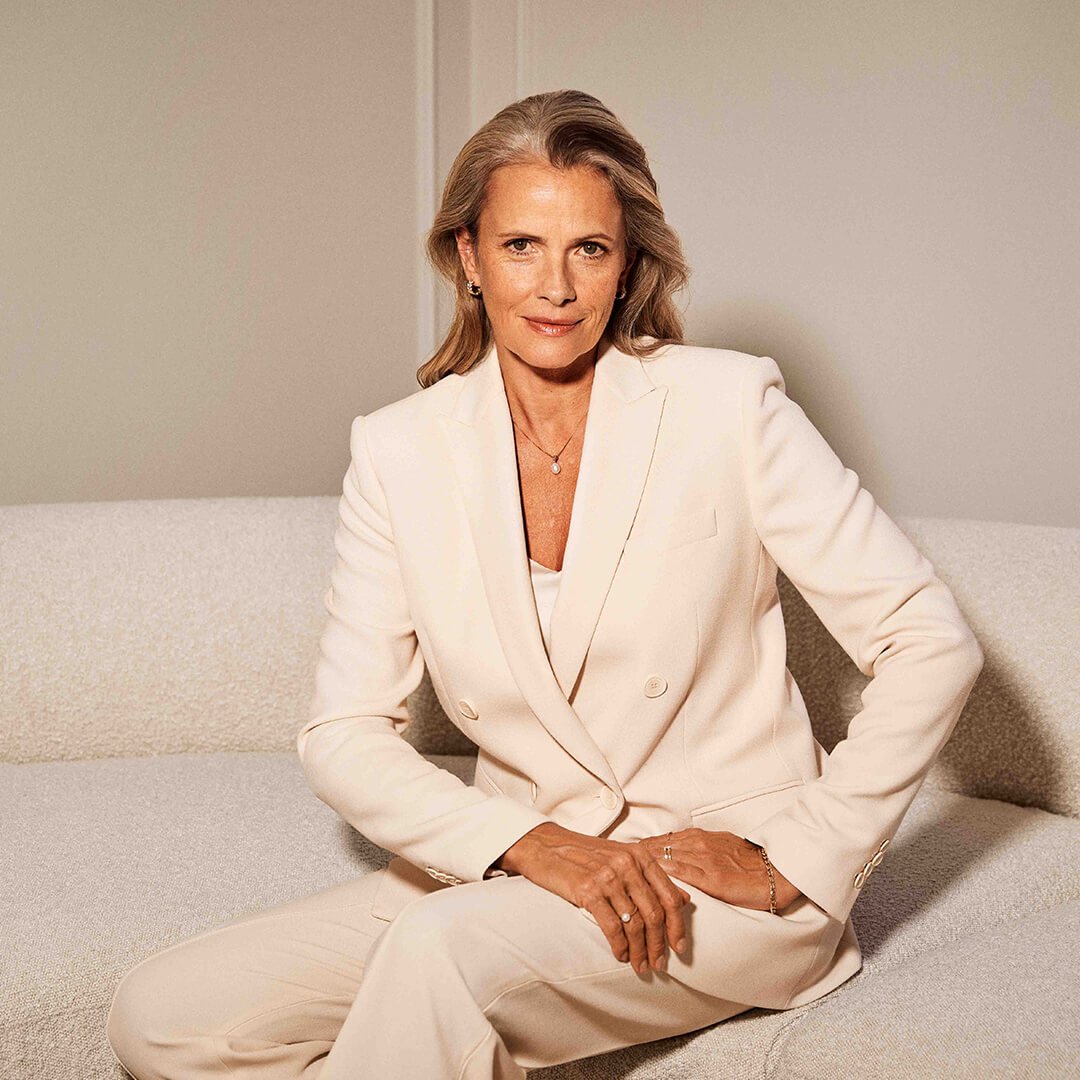 Voksen, smilende kvinne med grått hår i en kremhvit dress, sittende i en lys sofa. Hun har på seg elegante gullsmykker."