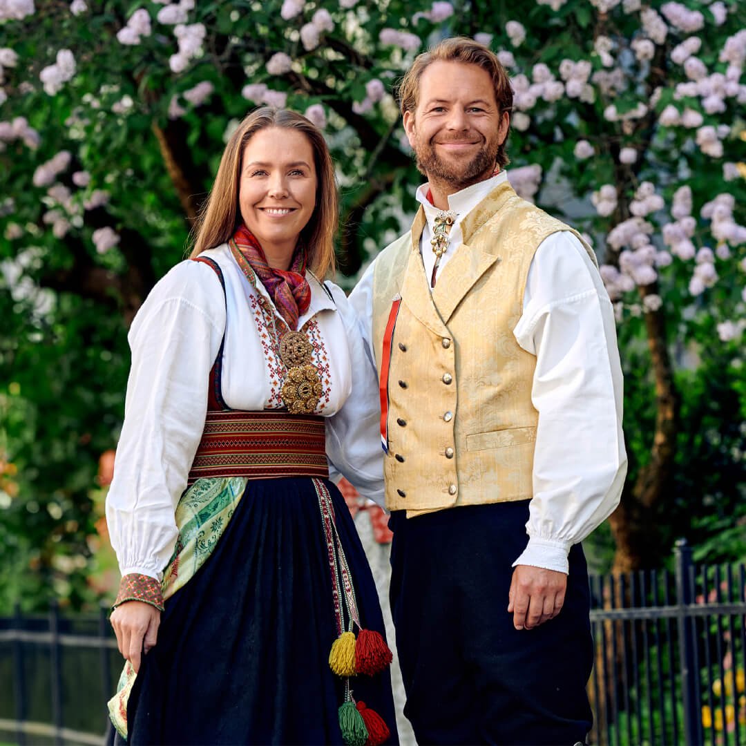 To personer i bunad står ute foran et tre med blomster, en kvinne i mørk bunad og en mann i lys vest.