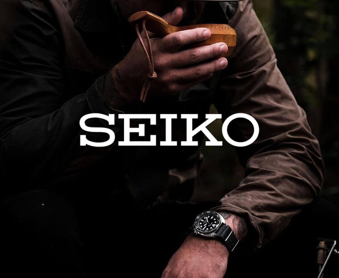 Mann i turklær med mørk Seiko-klokke på håndleddet drikker av trekopp i skogen, med SEIKO-logo sentrert i bildet.