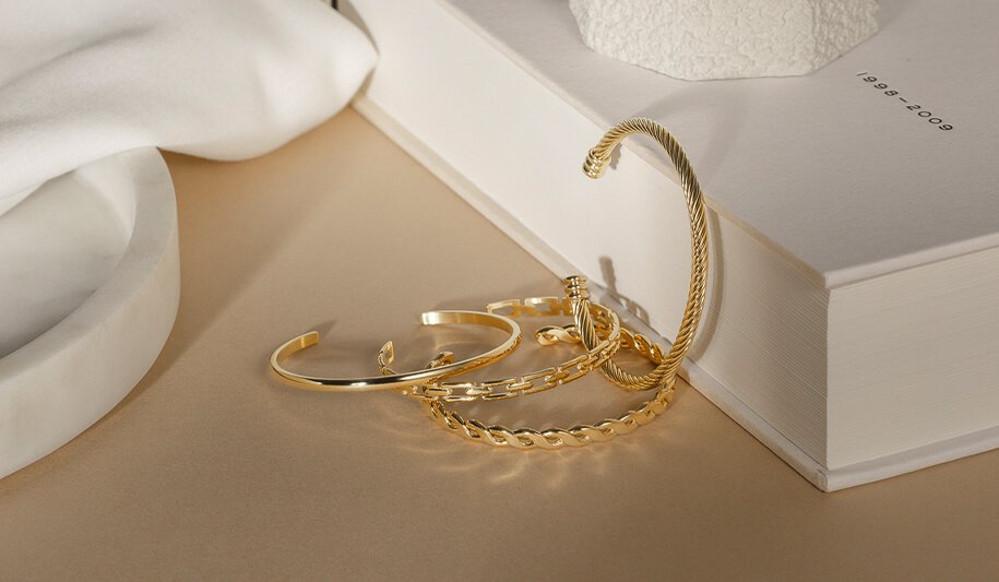 Fire armbånd i gult metall med ulike design ligger på et beige underlag ved siden av en bok og et hvitt stoff.