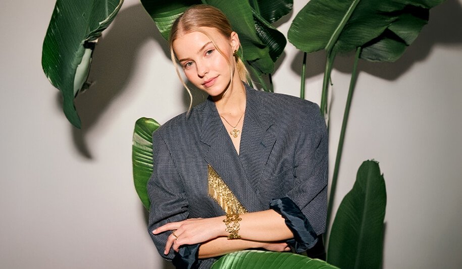 Kvinne foran grønne blader iført grå blazer med gullsmykker, armbånd og halskjede med blomstermotiv. 