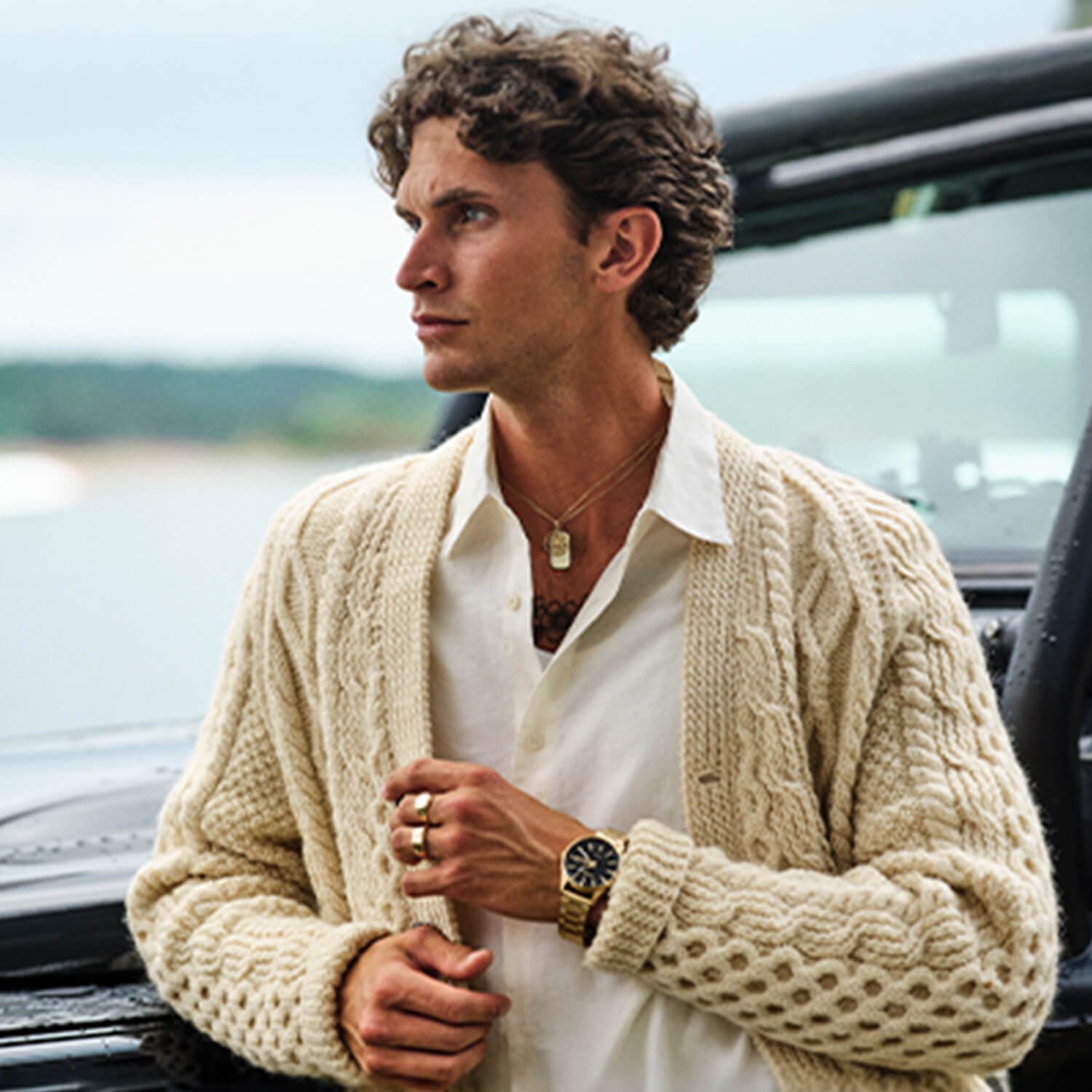 Mann med krøllete hår iført hvit skjorte og strikket beige cardigan, med klokke, ringer og halskjeder, står ved svart bil med åpent vindu.