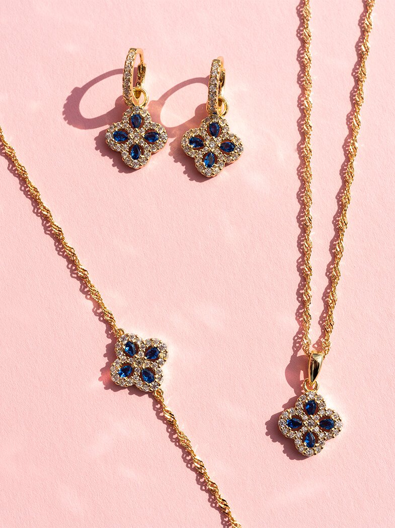 Gullsmykkesett med øredobber, armbånd og kjede med mørkeblå og klare steiner i kløverform på rosa bakgrunn.