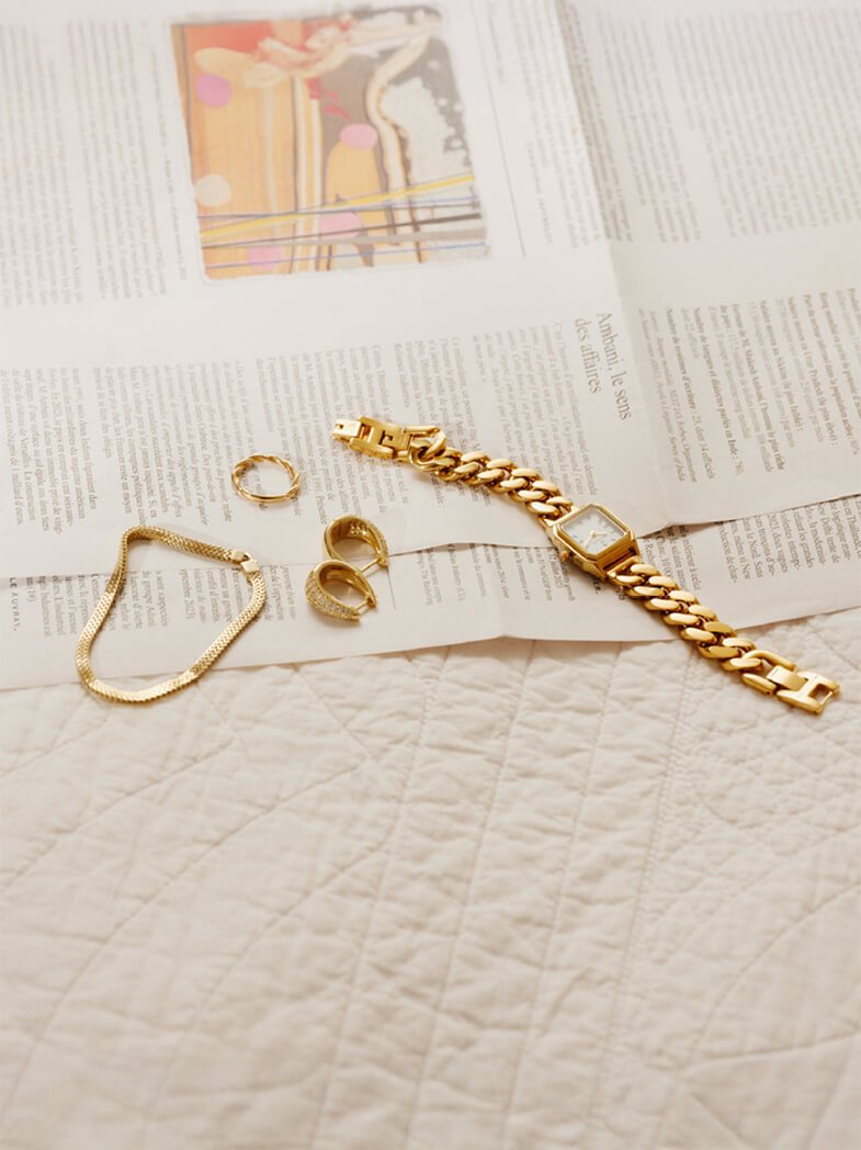 Ulike gullsmykker og en klokke ligger spredt utover en avis på en hvit, teksturert flate. Bildet har et varmt lys.