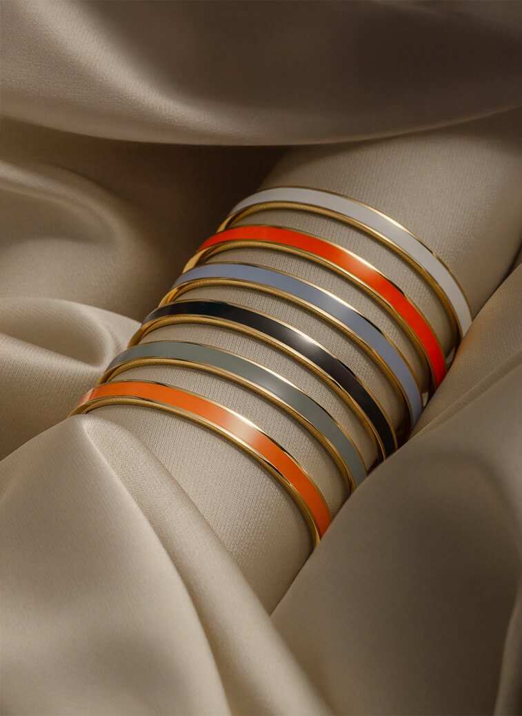 Fargerike gullbelagte armbånd i oransje, blå, grå og hvite nyanser ligger stablet på beige silkestoff.
