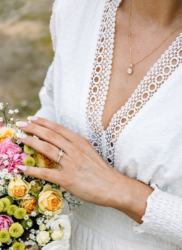 En kvinne ute på gress i hvit sommerkjole med matchende ring og kjede med anheng, og som holder en blomsterbukett i ene hånden.