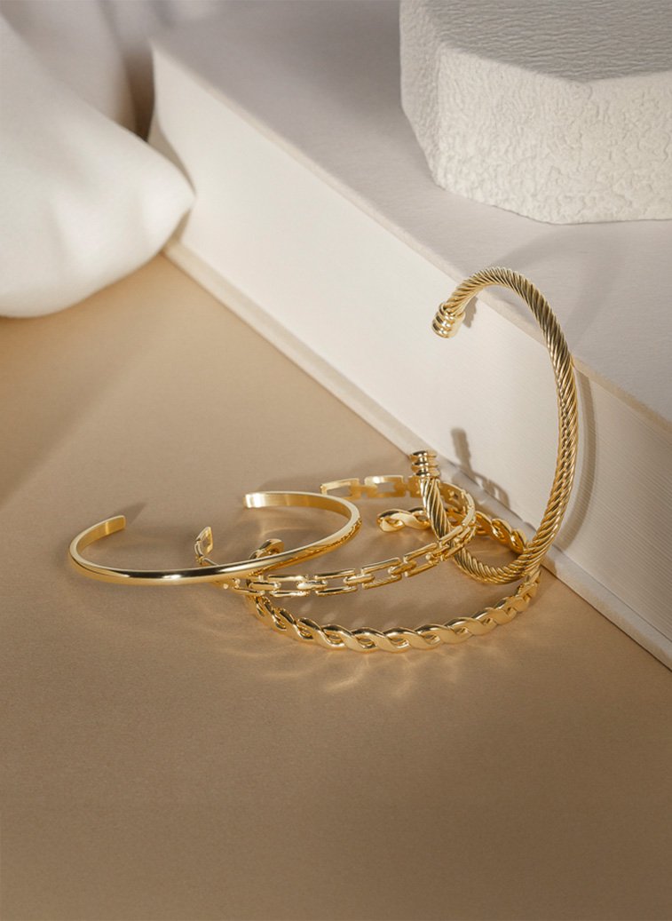 Fire gullfargede armbånd i ulike design ligger på beige overflate ved siden av en hvit bok og dekorstein.