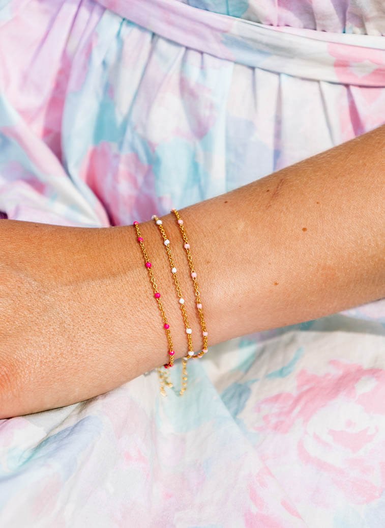 Armen til en sommerbrun kvinne i sommerkjole, med tre armbånd i samme gullfarge som har mørkerosa, hvit og lyserosa stener på.