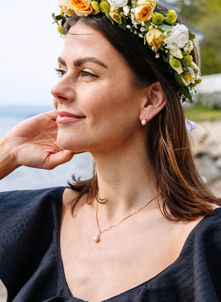 En kvinne med blomsterkrans på hodet, perleøredobber, kjede med perleanheng og sort kjole som smiler fornøyd og ser utover fjorden.