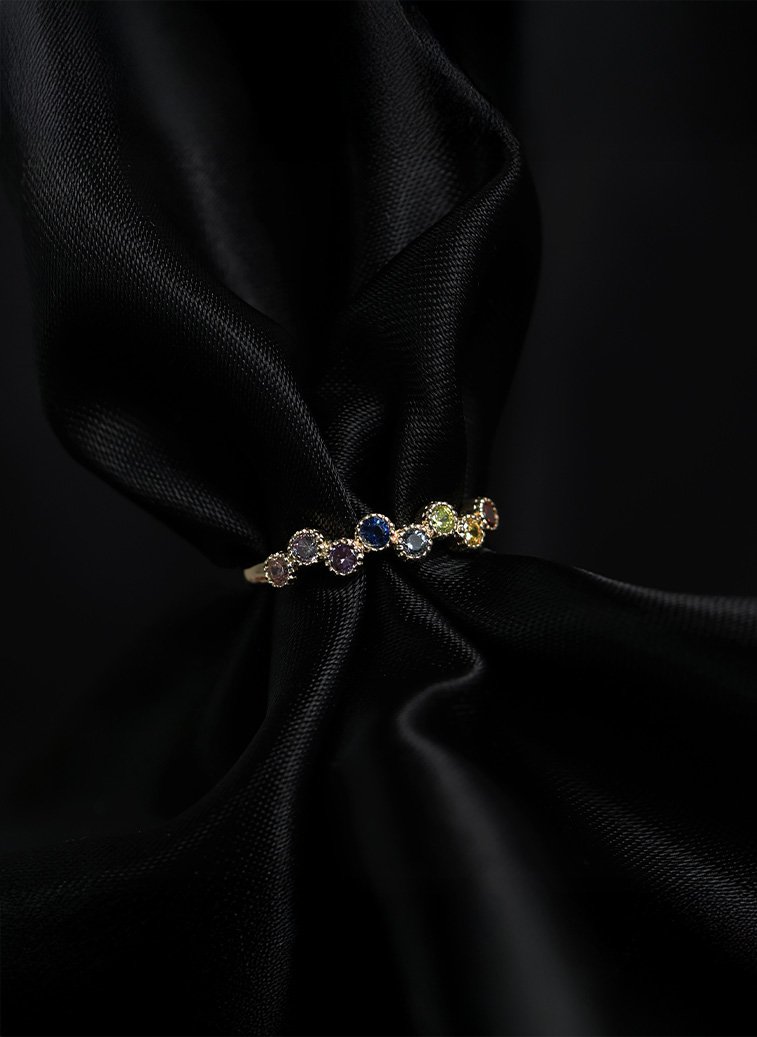 Gullring med runde fargede stener i blå, lilla, grønn og gul nyanse, plassert på sort silkestoff.