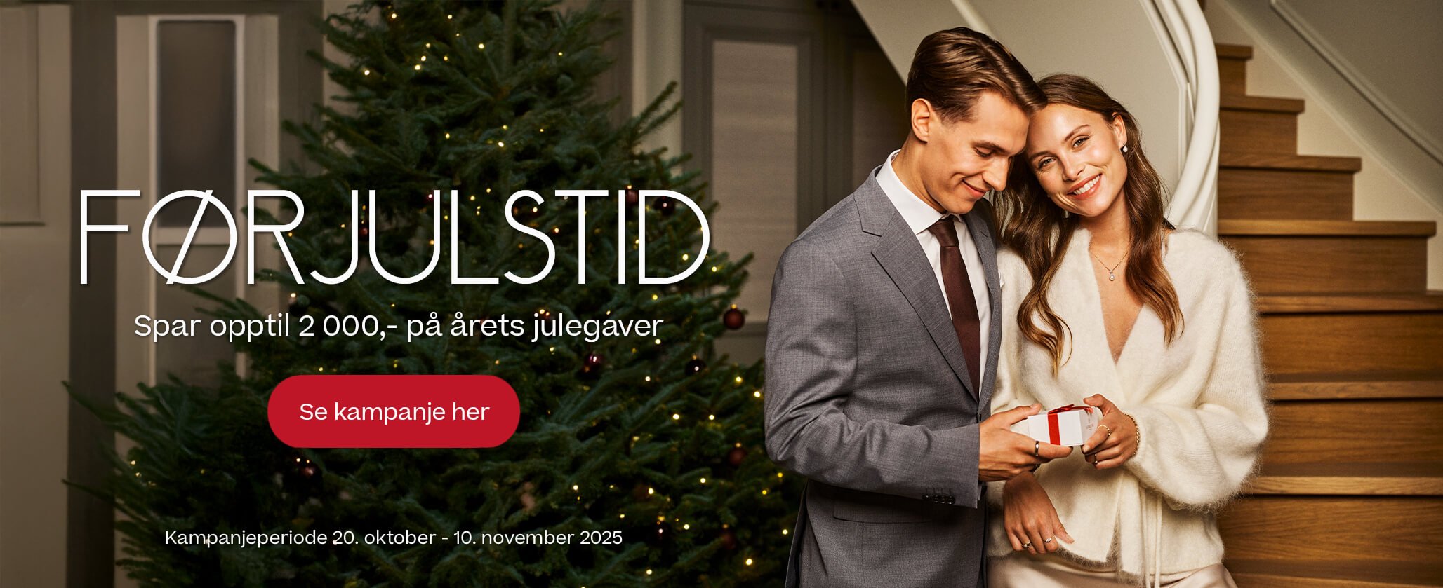 Par i elegant antrekk holder rød gaveeske foran pyntet juletre i moderne interiør. Mann i grå dress og kvinne i hvit genser med smykker.