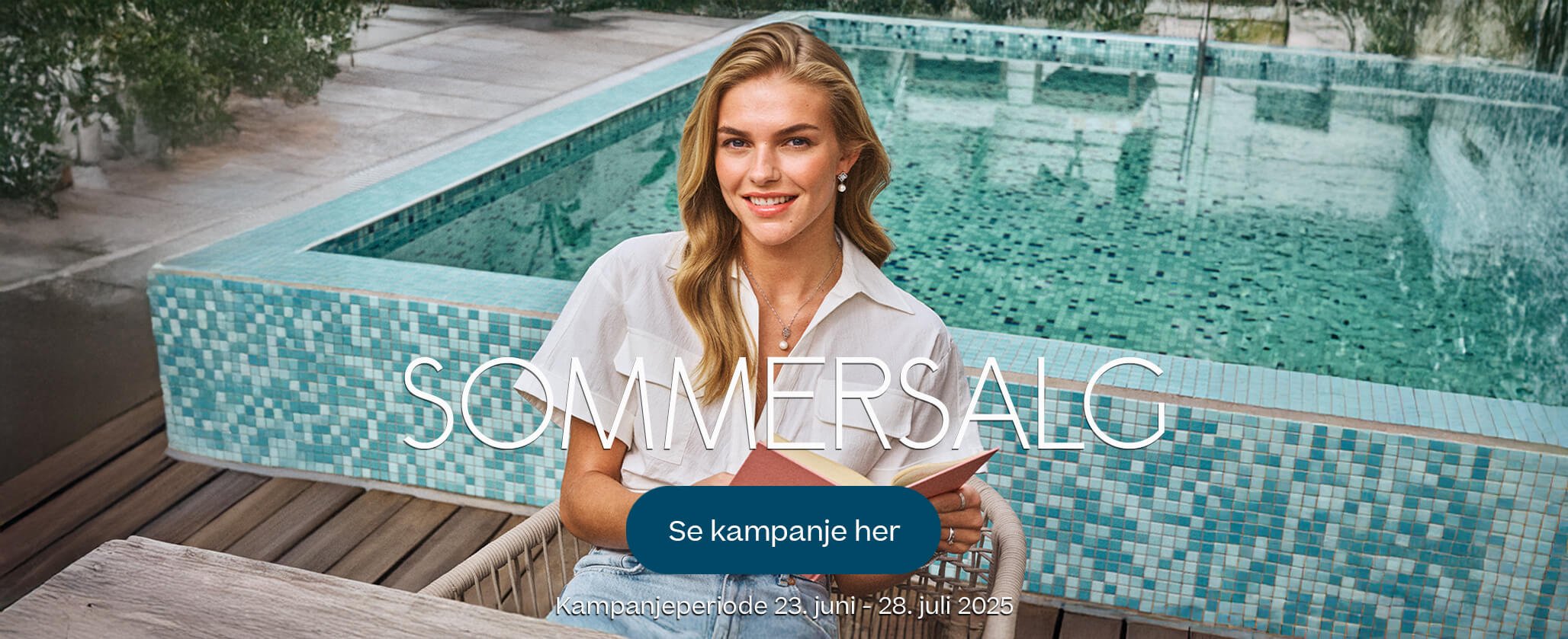 Kvinne med langt blondt hår sitter foran et basseng, iført hvit skjorte og smykker, mens hun holder en bok.