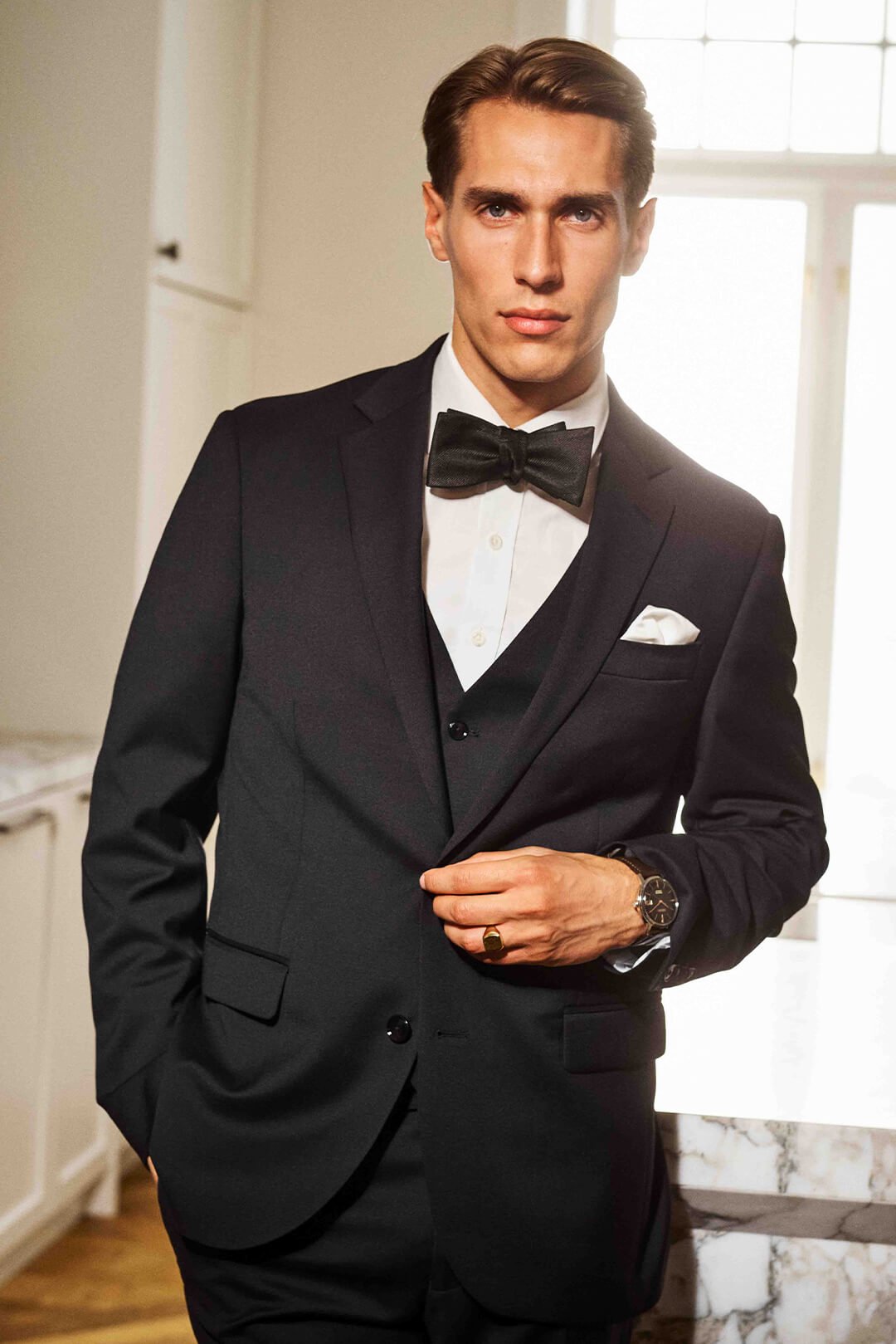 Mann i smoking med sløyfe, hvit skjorte og lommetørkle bærer ring og klokke med mørk urskive. Elegant interiør med store vinduer i bakgrunnen.