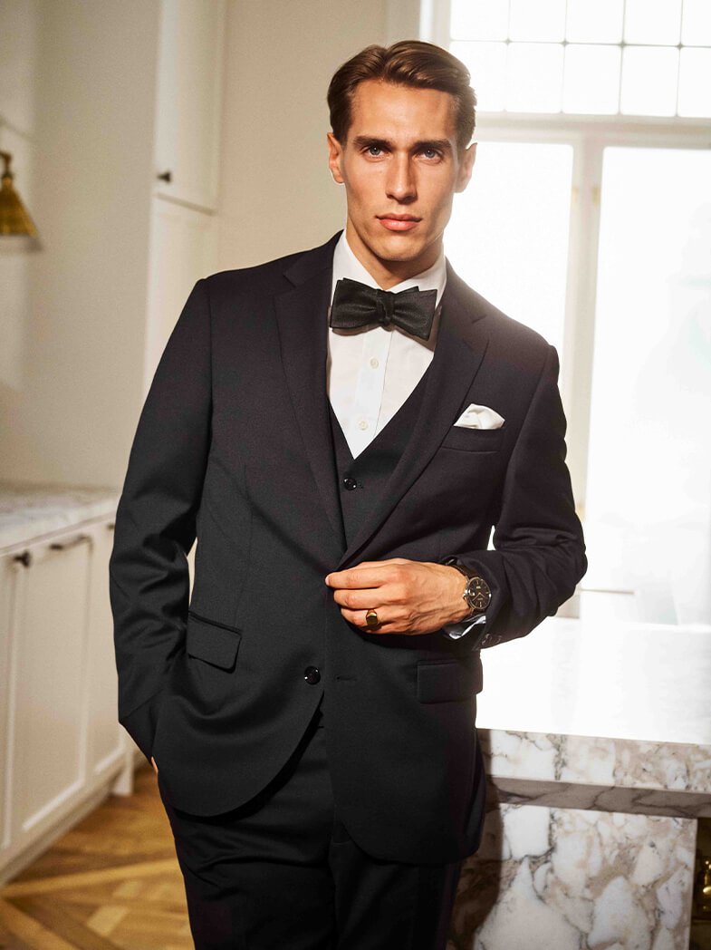 Mann i smoking med sløyfe, hvit skjorte og lommetørkle bærer ring og klokke med mørk urskive. Elegant interiør med store vinduer i bakgrunnen.