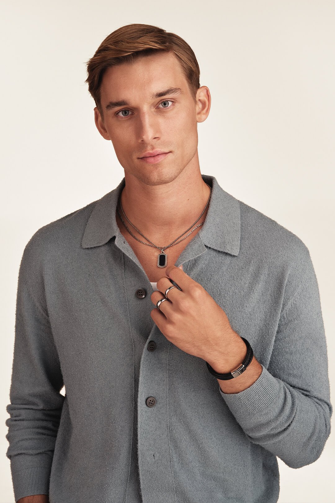 En ung mann med grå cardigan, skinn armbånd og halskjede, ser rett inn i kamera foran beige bakgrunn