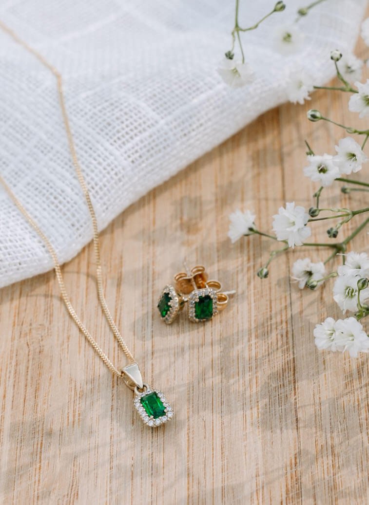 Et par gullbelagte øreringer og kjede med grønt smykke med små Cubic zirkonia, på et trebord med hvit duk og hviteblomster til venstre i bildet.