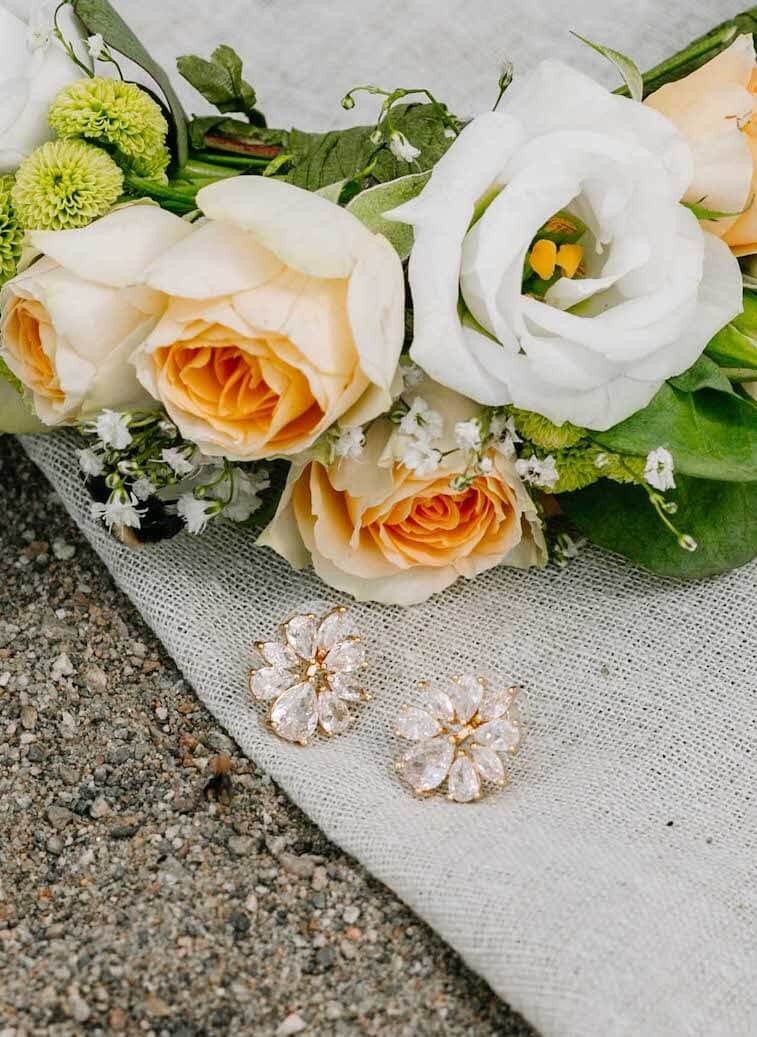 Et par hvite blomster øredobber som ligger på en hvit duk under en bukett med hvite roser og andre sommer blomster, på grusbakken.