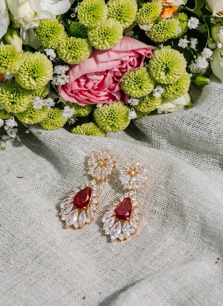 Et par øredobber i gullfarget metall, dekorert med fargerike cubic zirkonia stener, som ligger på en grå duk under en bukett med sommerblomster.