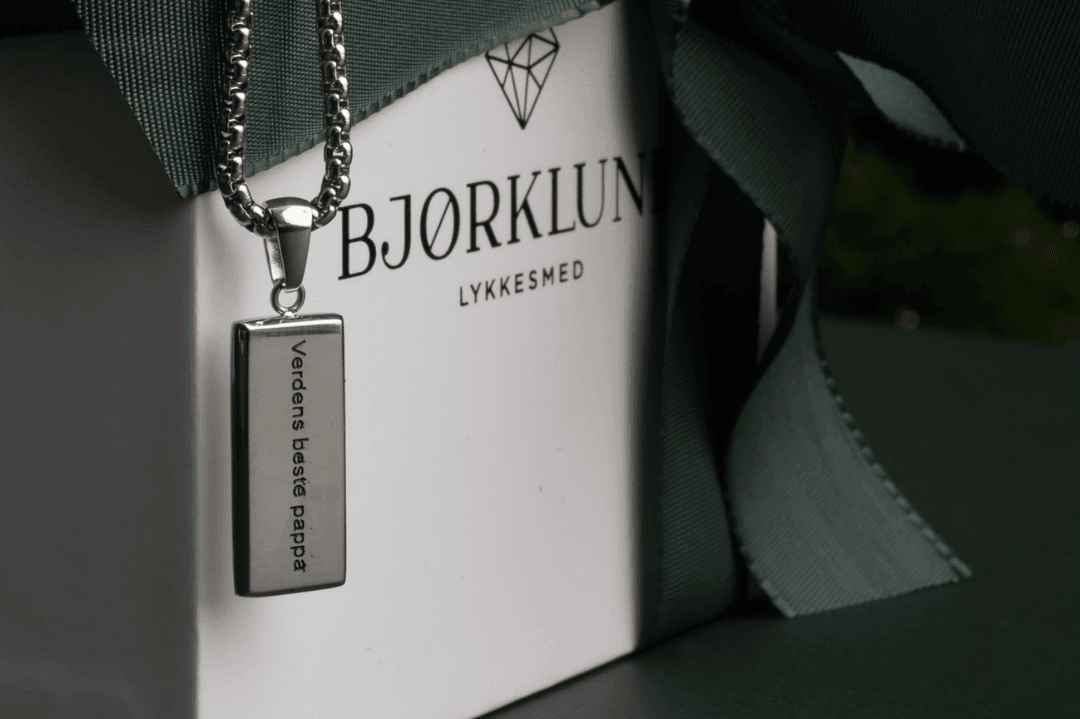 Verdens beste pappa-smykke fra Bjørklund Næbilde av smykke med tekst "verdens beste pappa" som ligger over en gaveboks fra Bjørklund